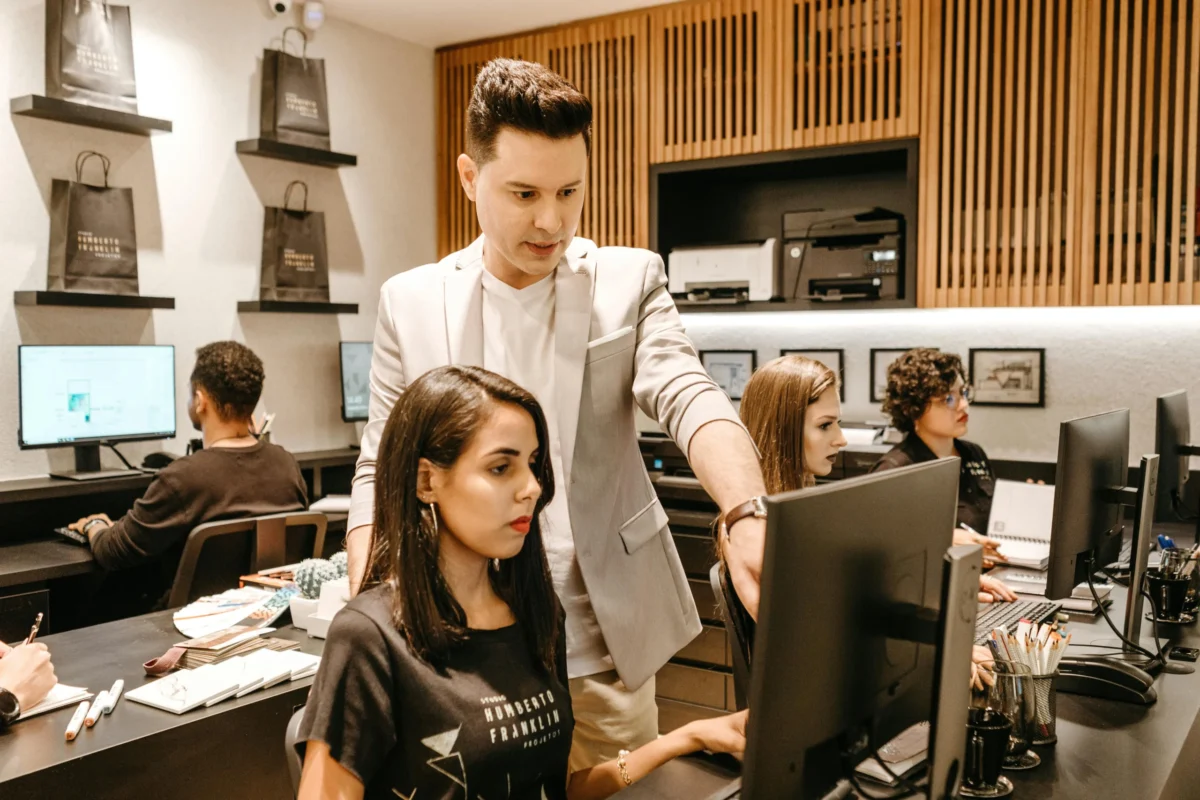 A colleague trains another on a computer in a busy office.