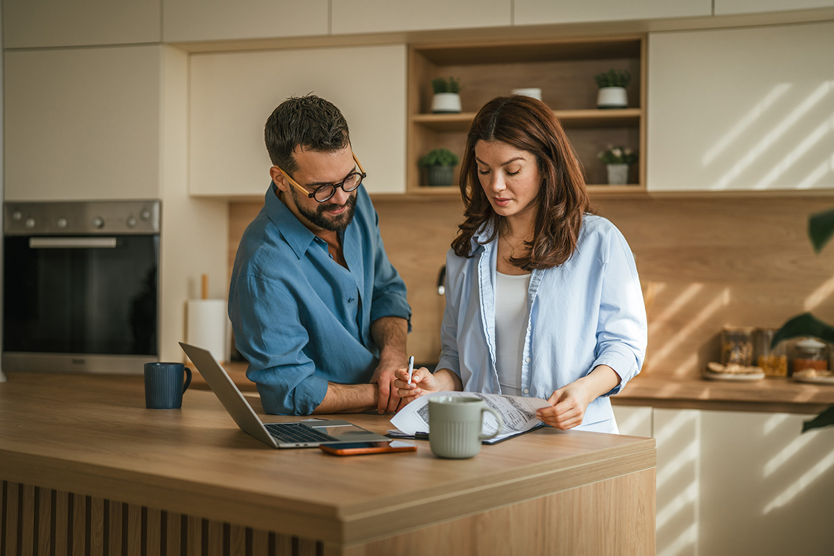 Couple looking at laptop and financial documents together in their kitchen.