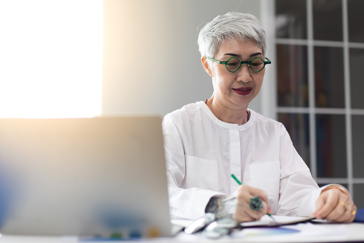 A mature business woman taking notes while working on a laptop.